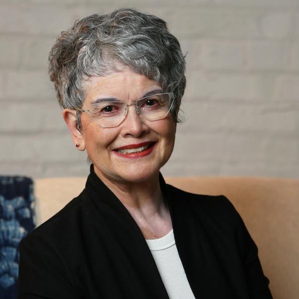 A person with short gray curly hair and glasses is smiling at the camera. They're wearing a black blazer or cardigan over a white top and red lipstick.